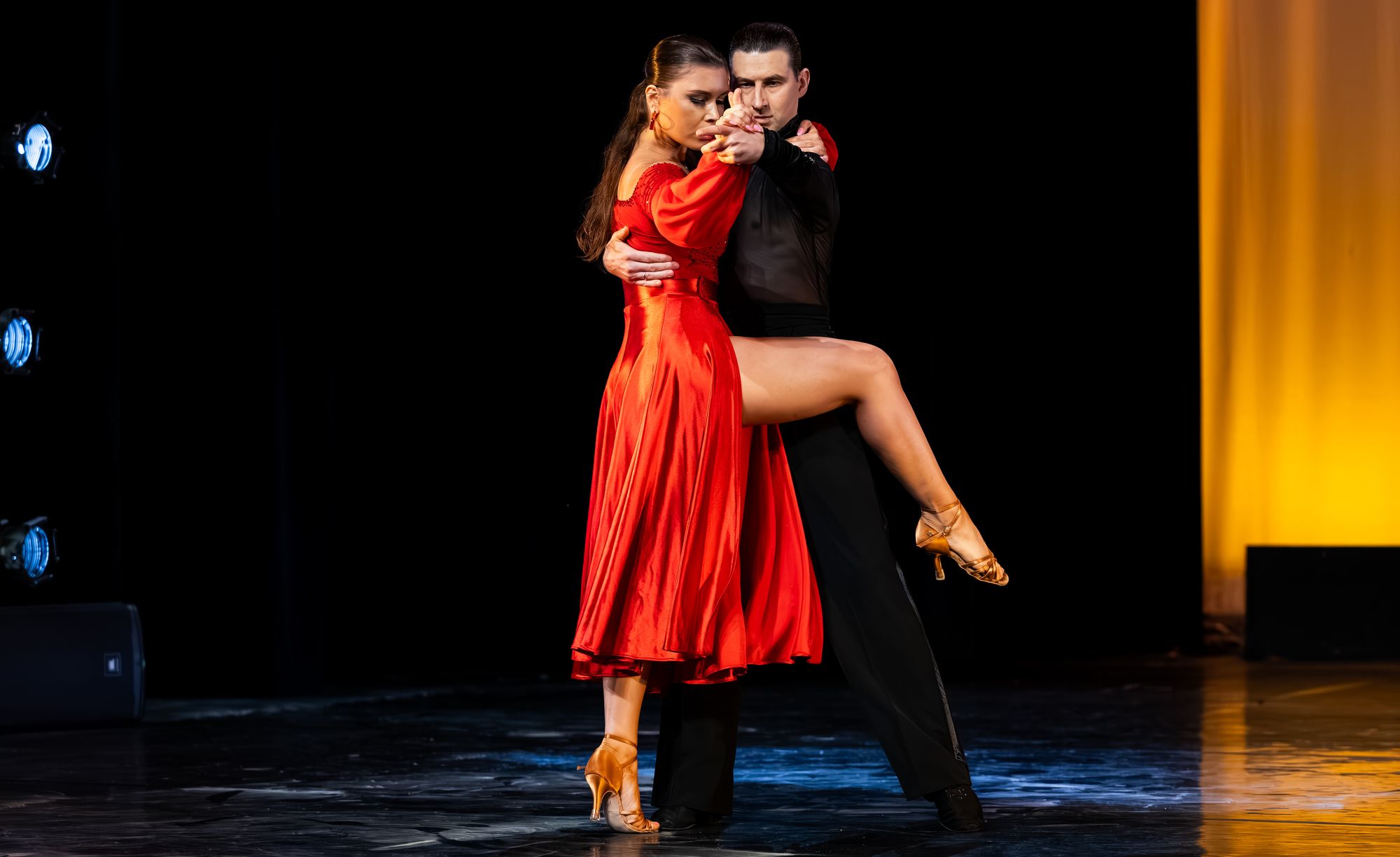 Two championship ballroom dancers hold a tango pose in a spotlight on the darkened Lewis Auditorium stage, the woman in a long red dress.