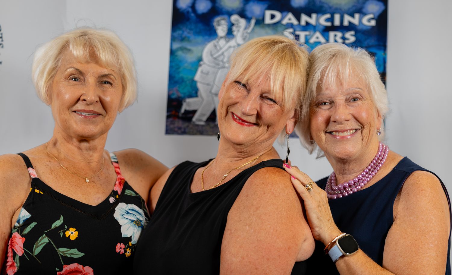 Three women smiling together in front of a Dancing Stars of St. Johns County poster at the 2026 show.